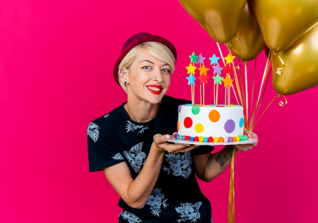 Sorridente ragazza bionda giovane partito indossando il cappello del partito che tiene palloncini e torta di compleanno con le stelle che guarda l'obbiettivo isolato su sfondo cremisi con lo spazio della copia
