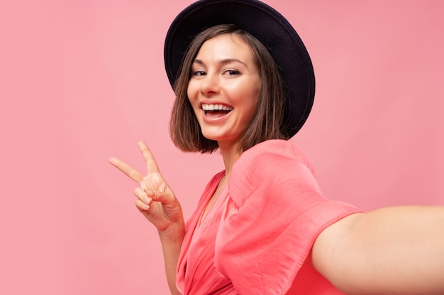 Sorridente ragazza bruna che fa autoritratto e posa sul muro rosa.