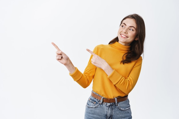 Sorridente ragazza bruna che punta le dita a sinistra e fissa l'affare promozionale, mostrando uno spazio vuoto pubblicitario, in piedi sul muro bianco