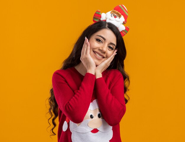 Sorridente ragazza carina che indossa la fascia di babbo natale e un maglione che tiene le mani sul viso isolato sul muro arancione con spazio di copia