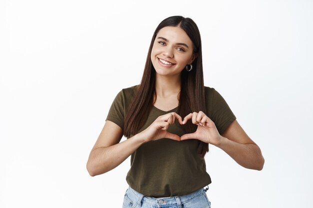 Sorridente ragazza carina che mostra il gesto del cuore sul petto e sembra romantica davanti, innamorandosi, come te, in piedi sul muro bianco