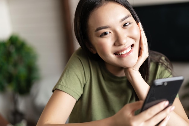 Sorridente ragazza coreana che sembra felice tenendo lo smartphone e seduta in salotto a casa lifestyle co...