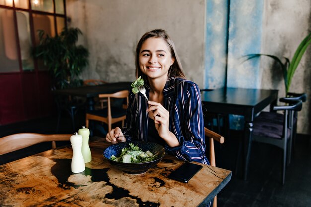 Sorridente ragazza dagli occhi scuri che mangia insalata nella caffetteria. Modello femminile allegro che gode del piatto vegetariano.