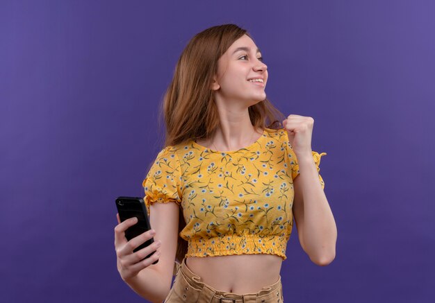 Sorridente ragazza giovane azienda telefono cellulare e alzando il pugno guardando il lato destro sulla parete viola isolata