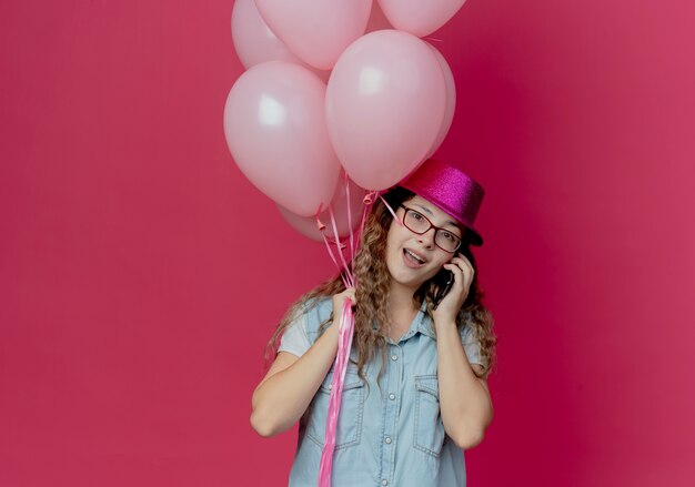 Sorridente ragazza giovane con gli occhiali e cappello rosa tenendo palloncini e parla al telefono isolato in rosa