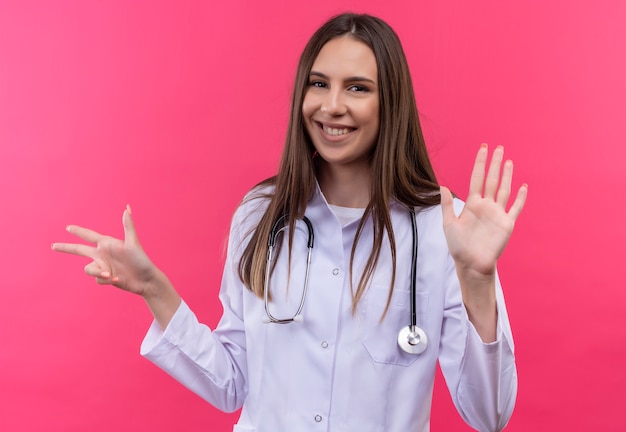 Sorridente ragazza giovane medico indossando stetoscopio abito medico che mostra numeri diversi su sfondo rosa isolato