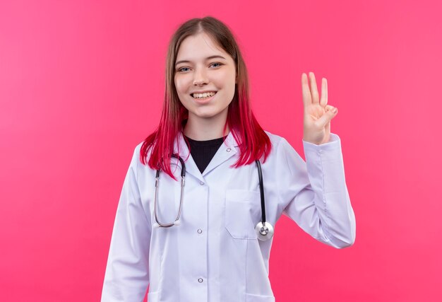 Sorridente ragazza giovane medico indossando stetoscopio abito medico che mostra tre su sfondo rosa isolato