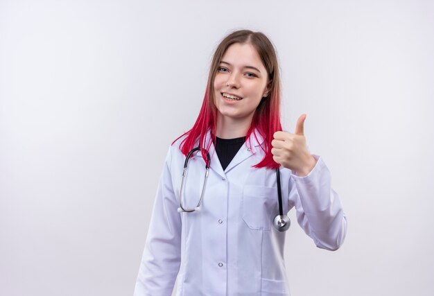 Sorridente ragazza giovane medico indossando stetoscopio abito medico il pollice in alto su sfondo bianco isolato