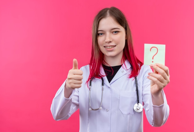 Sorridente ragazza giovane medico indossando stetoscopio abito medico tenendo la carta punto interrogativo il suo pollice in alto su sfondo rosa isolato