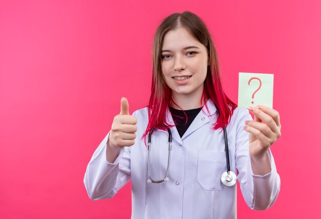 Sorridente ragazza giovane medico indossando stetoscopio abito medico tenendo la carta punto interrogativo il suo pollice in alto su sfondo rosa isolato