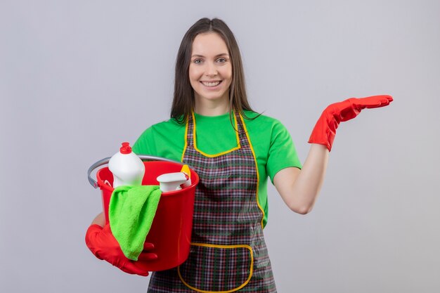 Sorridente ragazza giovane pulizia indossando uniformi in guanti rossi in possesso di strumenti di pulizia alzando la mano su sfondo bianco isolato