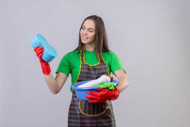 Sorridente ragazza giovane pulizia indossando uniformi in guanti rossi in possesso di strumenti per la pulizia guardando la spugna sulla sua mano su sfondo bianco isolato