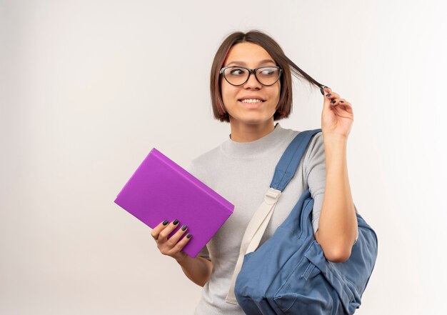 Sorridente ragazza giovane studente con gli occhiali e borsa posteriore che tiene il libro e i suoi capelli guardando il lato isolato sul muro bianco