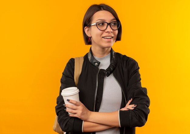 Sorridente ragazza giovane studente con gli occhiali e borsa posteriore in piedi con la postura chiusa tenendo la tazza di caffè in plastica guardando il lato isolato sulla parete arancione