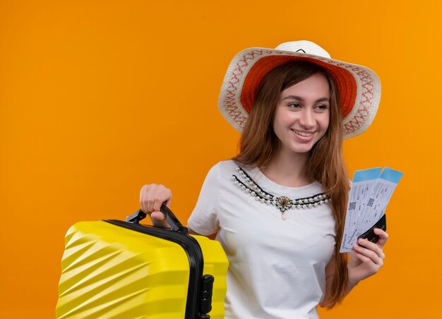Sorridente ragazza giovane viaggiatore che indossa il cappello che tiene la valigia e biglietti aerei, carta di credito sulla parete arancione isolata