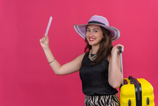 Sorridente ragazza giovane viaggiatore che indossa la maglietta nera in cappello tenendo la valigia e biglietti su sfondo rosso