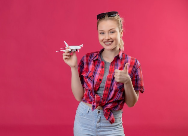 Sorridente ragazza giovane viaggiatore indossa una camicia rossa e bicchieri sulla sua testa tenendo il piano di giocattoli il suo pollice in alto su sfondo rosa isolato