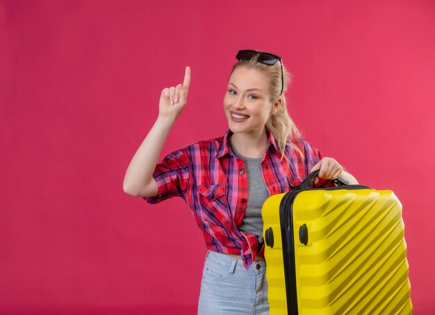 Sorridente ragazza giovane viaggiatore indossa una camicia rossa e occhiali sulla sua testa tenendo la valigia punta su sfondo rosa isolato