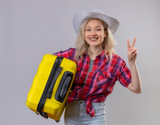 Sorridente ragazza giovane viaggiatore indossa una camicia rossa in cappello tenendo la valigia che mostra il gesto di pace isolato su sfondo bianco