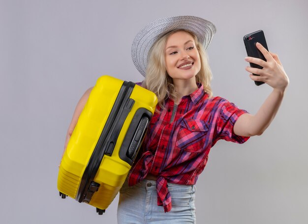 Sorridente ragazza giovane viaggiatore indossa una camicia rossa in cappello tenendo la valigia e tenendo selfie su sfondo bianco isolato