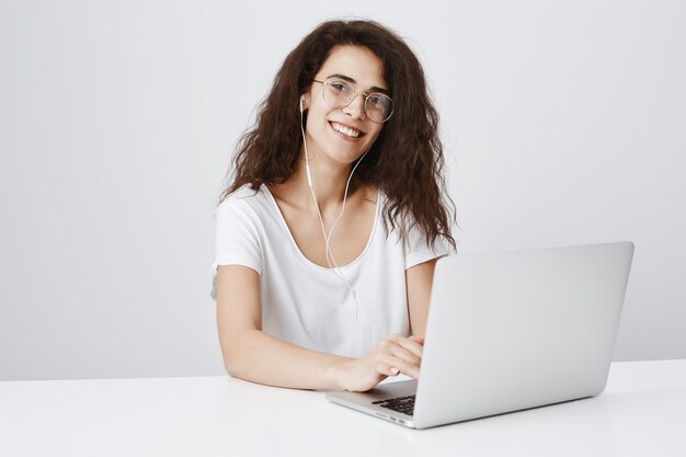 Sorridente ragazza graziosa ascoltando musica in auricolari, lavorando su laptop