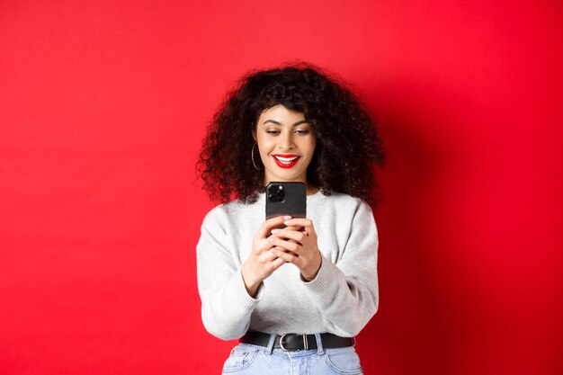 Sorridente ragazza moderna che scatta foto su smartphone, guarda lo schermo e registra video, in piedi su sfondo rosso