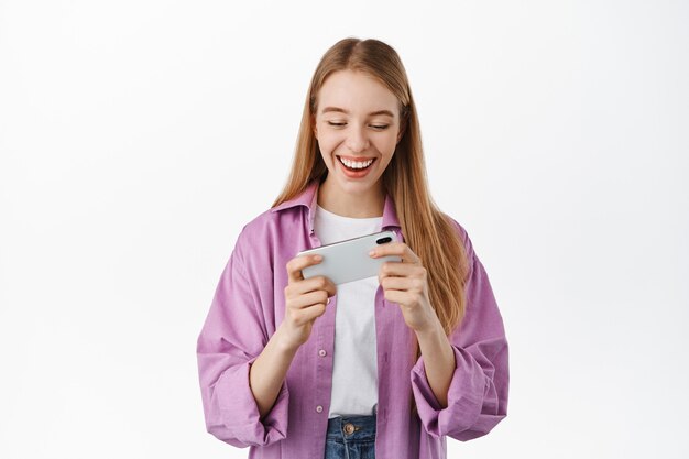 Sorridente ragazza moderna che tiene lo smartphone con entrambe le mani, guardando lo schermo mobile orizzontale, guardando i video sul telefono o giocando ai videogiochi, in piedi sul muro bianco