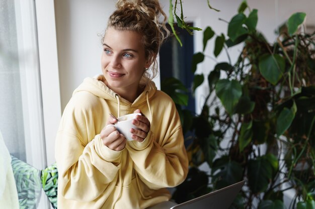 Sorridente ragazza sognante guardando fuori dalla finestra, bere caffè e lavorare a distanza da casa utilizzando il computer portatile.