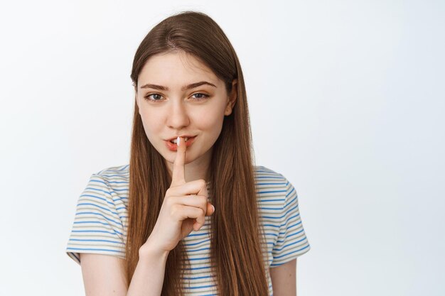 Sorridente ragazza timida zitta, premi il dito sulle labbra con il gesto shh shush, raccontando il segreto, stai tranquillo segno, in piedi su sfondo bianco