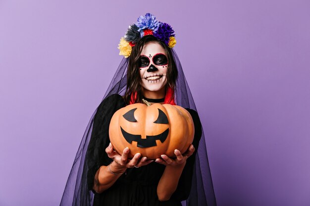 Sorridente ragazza zombie in velo nero in posa sulla parete pastello. Felice donna in abito da sposa morta che tiene la zucca di Halloween.