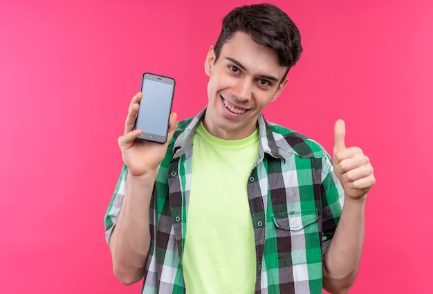 Sorridente ragazzo giovane caucasico indossa una camicia verde tenendo il telefono con il pollice in alto su sfondo rosa isolato