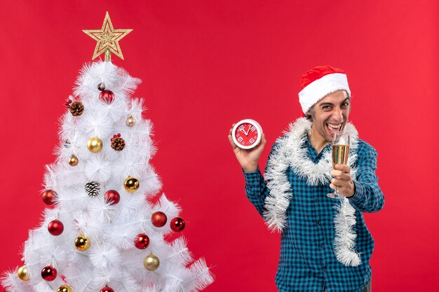 Sorridente ragazzo giovane divertente con cappello di Babbo Natale e alzando un bicchiere di vino e tenendo l'orologio in piedi vicino all'albero di Natale sul rosso