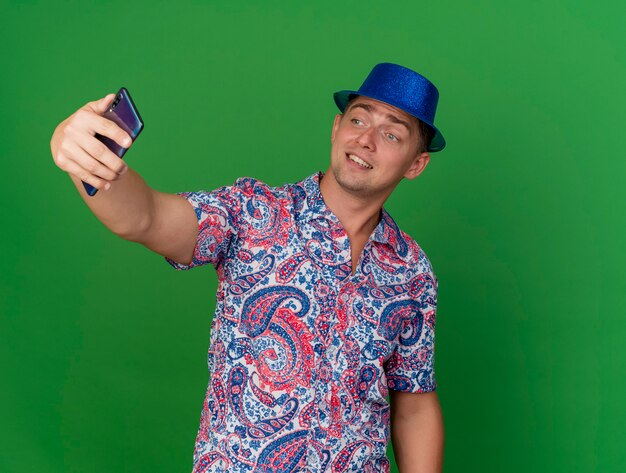 Sorridente ragazzo giovane partito che indossa cappello blu prendere un selfie isolato su sfondo verde