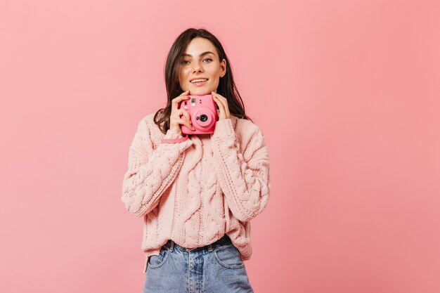 Sorridente signora dai capelli scuri in elegante maglione pone con fotocamera rosa su sfondo isolato.