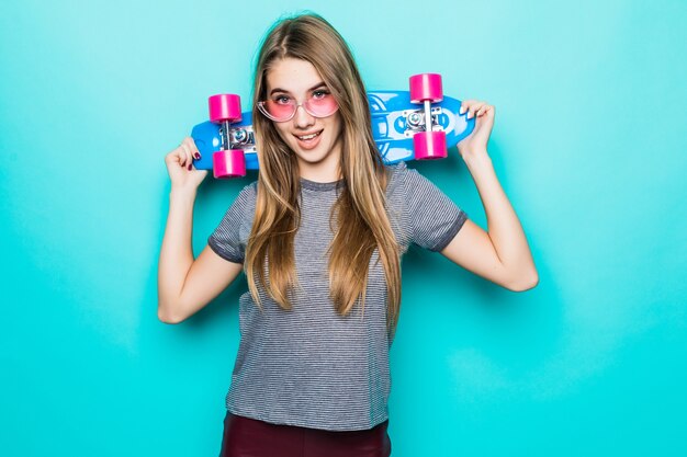 Sorridente skategirl adolescente dai capelli d'oro sta con skateboard colorato isolato su sfondo verde