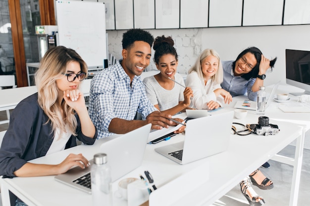 Sorridente studente africano che punta con la matita sullo schermo del laptop. Concentrato donna bionda in occhiali puntellando il mento con la mano mentre si lavora con il computer in ufficio.