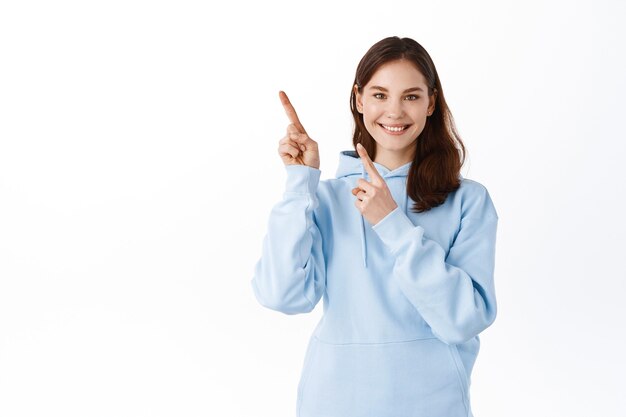 Sorridente studentessa carina che punta le dita da parte nell'angolo in alto a sinistra, mostrando pubblicità, in piedi in felpa con cappuccio contro il muro bianco