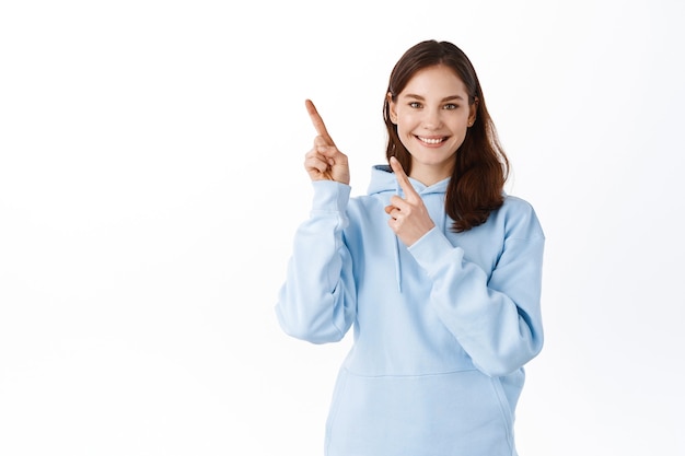 Sorridente studentessa carina che punta le dita da parte nell'angolo in alto a sinistra, mostrando pubblicità, in piedi in felpa con cappuccio contro il muro bianco