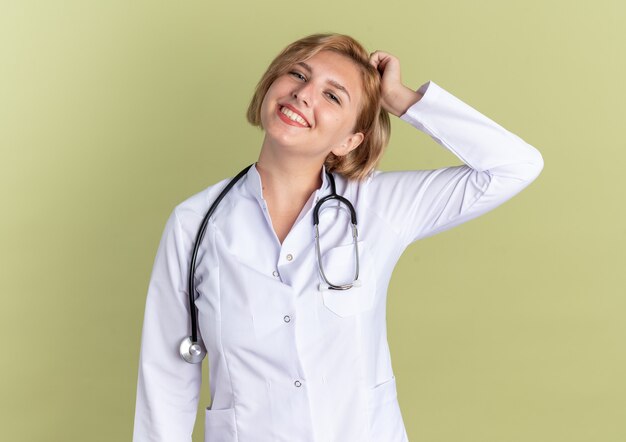 Sorridente testa inclinabile giovane dottoressa che indossa un abito medico con uno stetoscopio che mette la mano sulla testa isolata su sfondo verde oliva