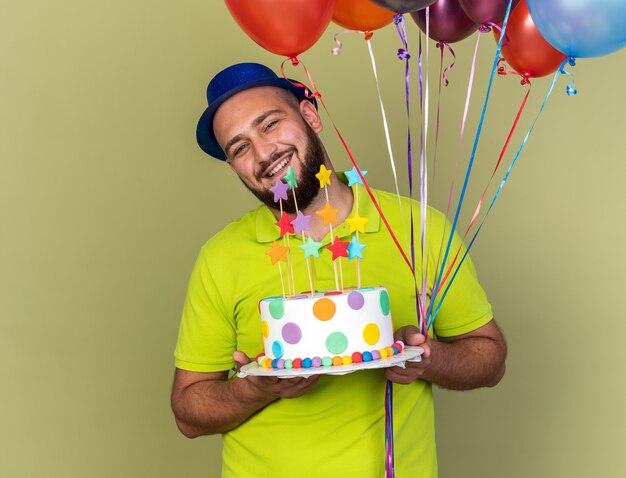 Sorridente testa inclinabile giovane uomo che indossa cappello da festa tenendo palloncini con torta