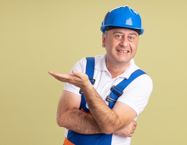 Sorridente uomo adulto costruttore in uniforme tiene la mano aperta su verde oliva