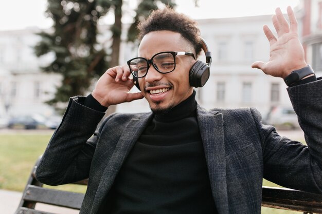 Sorridente uomo africano seduto sulla panchina ed esprimere emozioni positive. Ragazzo nero che ride ascoltando musica in cuffia al mattino.