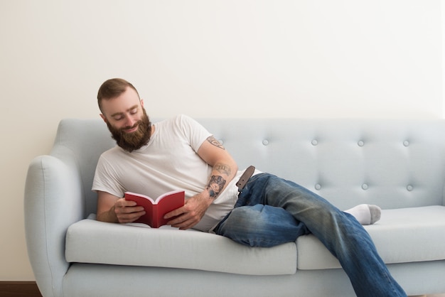 Sorridente uomo barbuto sdraiato sul divano e leggendo il libro