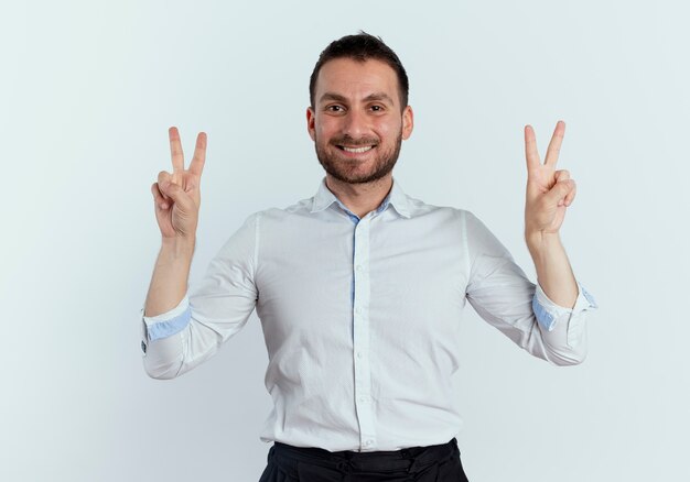 Sorridente uomo bello gesti il segno della mano di vittoria con due mani isolate sul muro bianco