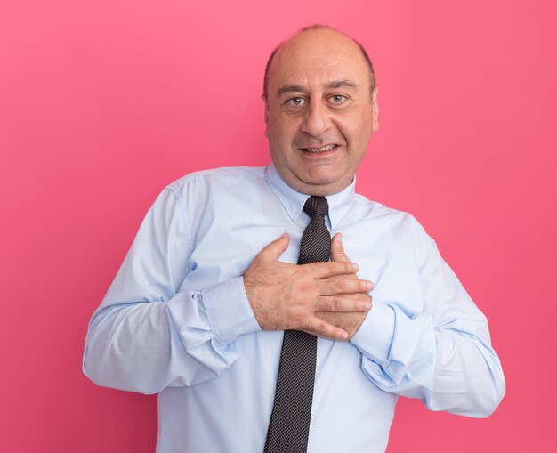 Sorridente uomo di mezza età che indossa la maglietta bianca con cravatta che tiene le mani sul cuore isolato sul muro rosa