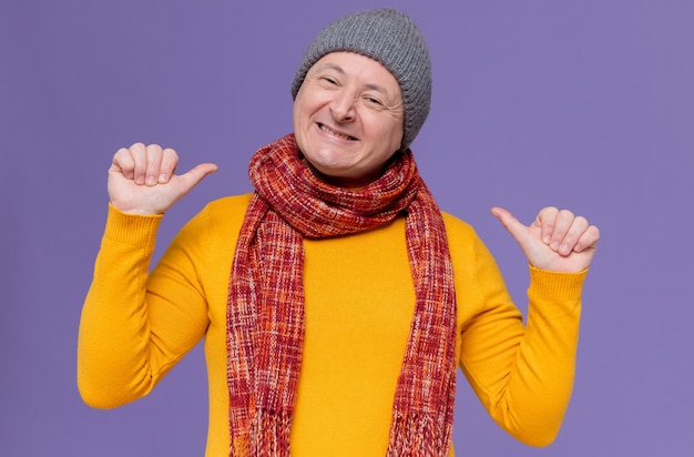 Sorridente uomo slavo adulto con cappello invernale e sciarpa intorno al collo che punta a se stesso