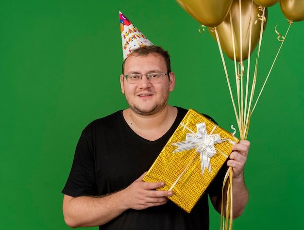 Sorridente uomo slavo adulto in occhiali ottici che indossa un berretto di compleanno tiene palloncini di elio e confezione regalo