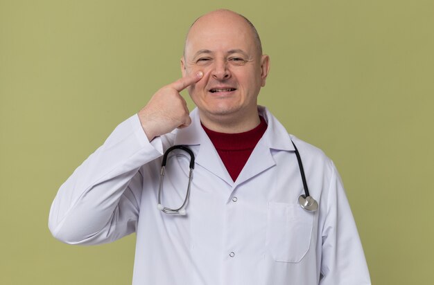 Sorridente uomo slavo adulto in uniforme da medico con stetoscopio che mette il dito sulla palpebra