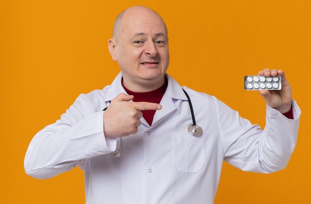 Sorridente uomo slavo adulto in uniforme da medico con stetoscopio tiene e indica il blister della medicina