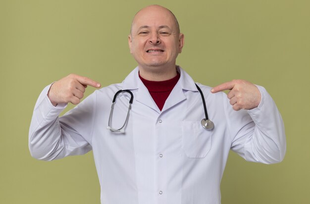 Sorridente uomo slavo adulto in uniforme da medico con uno stetoscopio che punta a se stesso